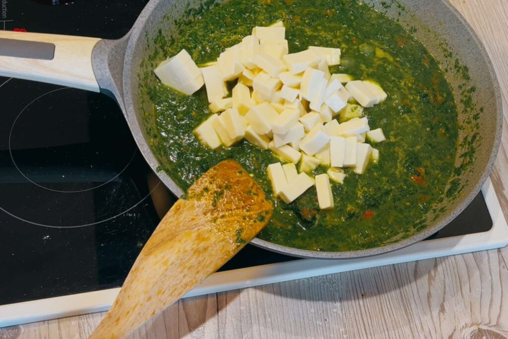 Frau fügt dem Spinat und den Gewürzen in der Pfanne das Paneer hinzu.